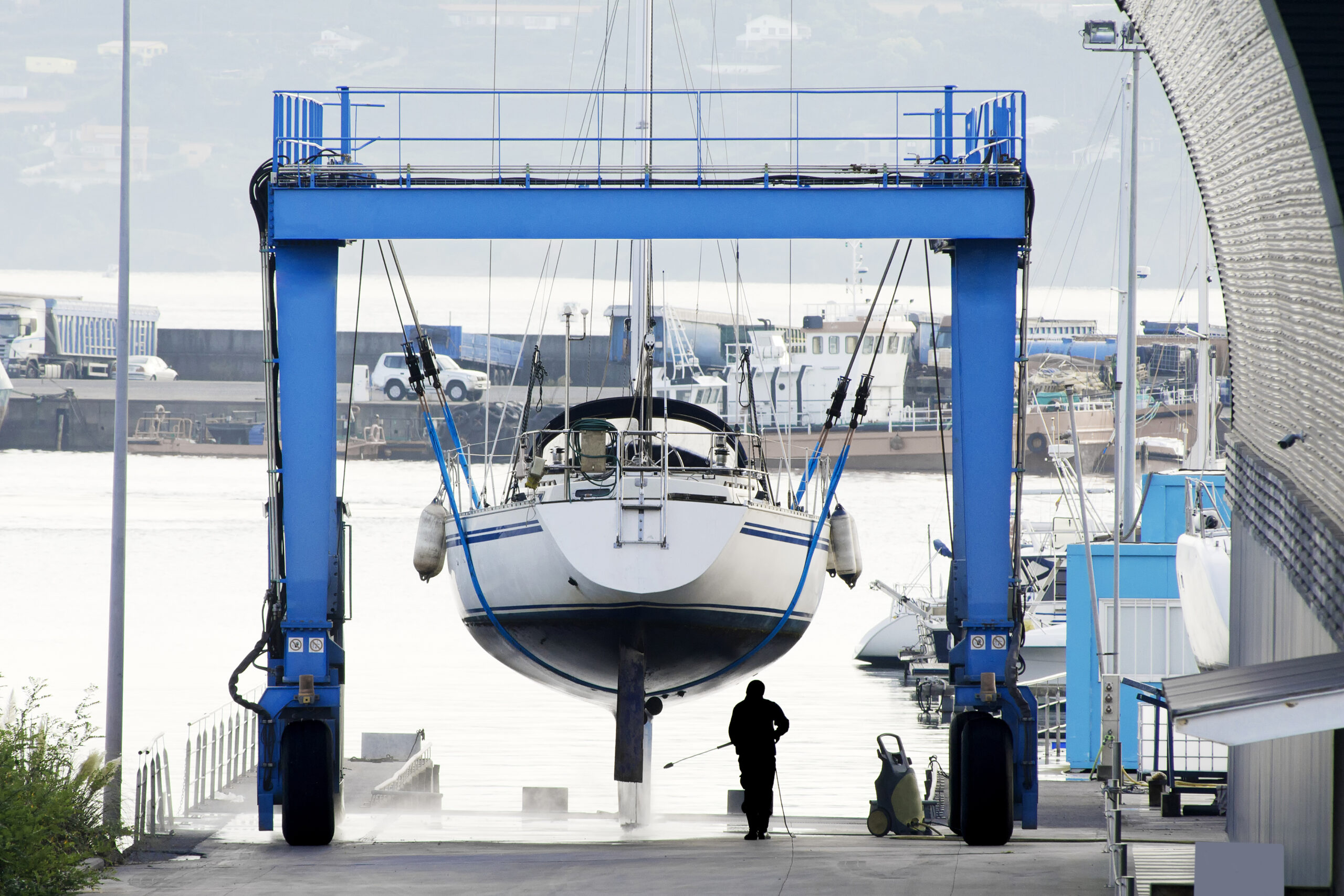 A sailboat is being lifted out of the water by a blue crane at a marina, with a silhouette of a worker cleaning nearby.