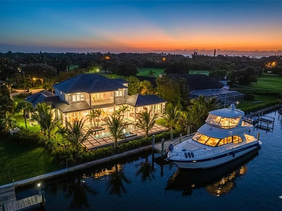 Luxurious waterfront home at twilight, featuring lush landscaping, a swimming pool, and a private yacht docked nearby, in a tranquil setting.
